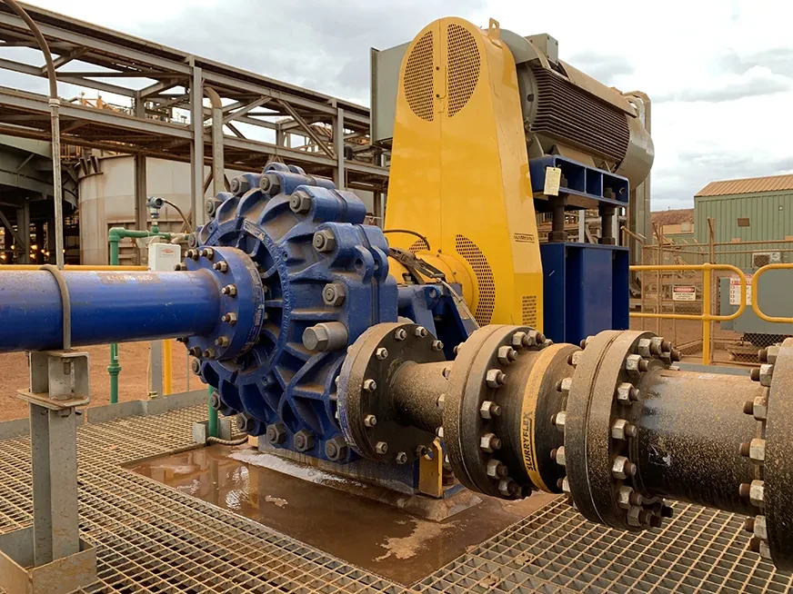 A slurry pump on a process plant: blue casing, flanged pipework and a yellow guarded motor on a steel grating platform